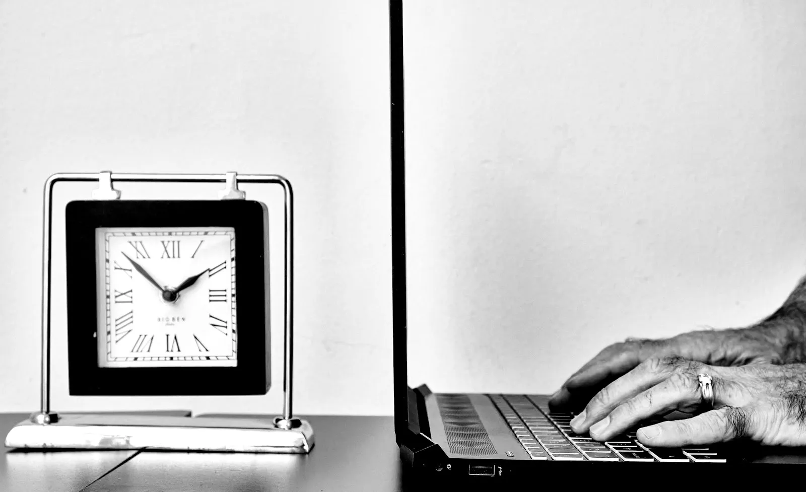 Iemand typt op een laptop aan een bureau, naast een grote wandklok